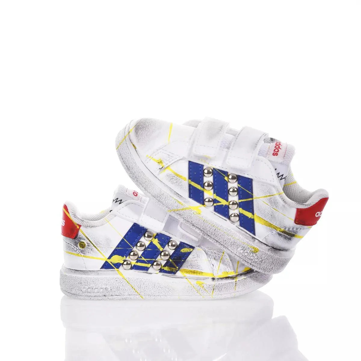 Scarpe da ginnastica bianche per bambini con dettagli blu, gialli e borchie, chiusura a strappo.