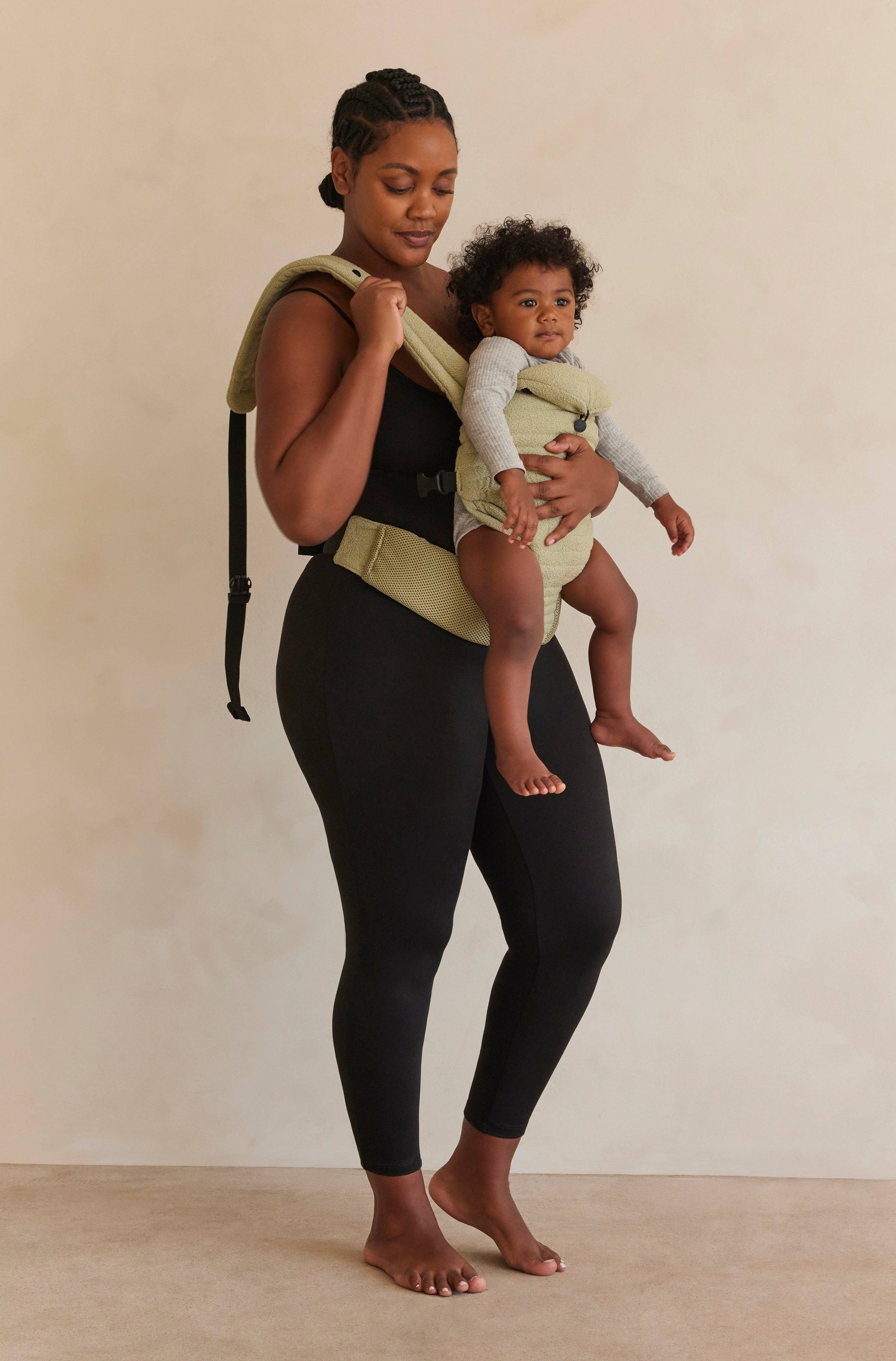 Donna indossa abbigliamento premaman nero e porta un bambino in marsupio beige