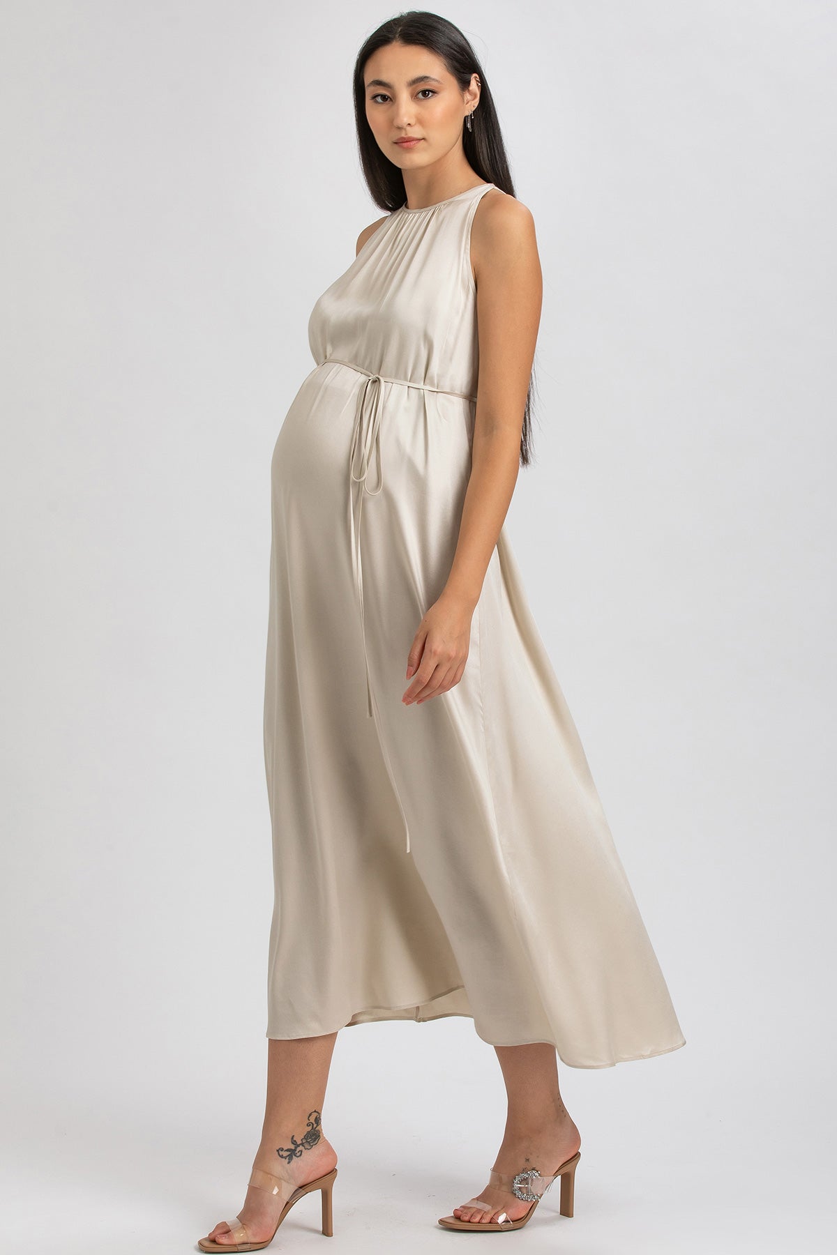 Donna incinta con abito premaman elegante beige in raso, sandali con tacco, fondo neutro