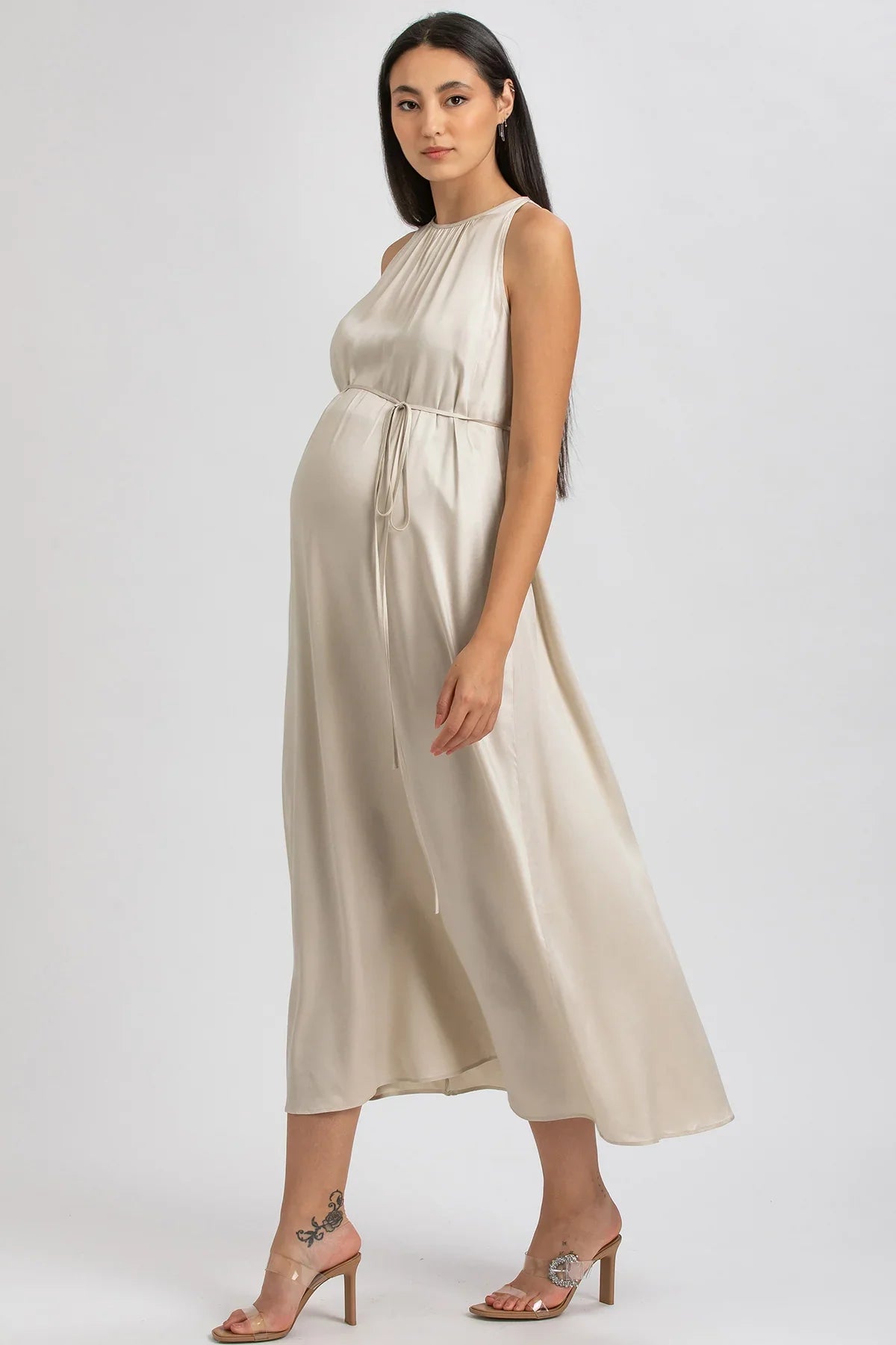Donna incinta con abito premaman elegante beige in raso, sandali con tacco, fondo neutro