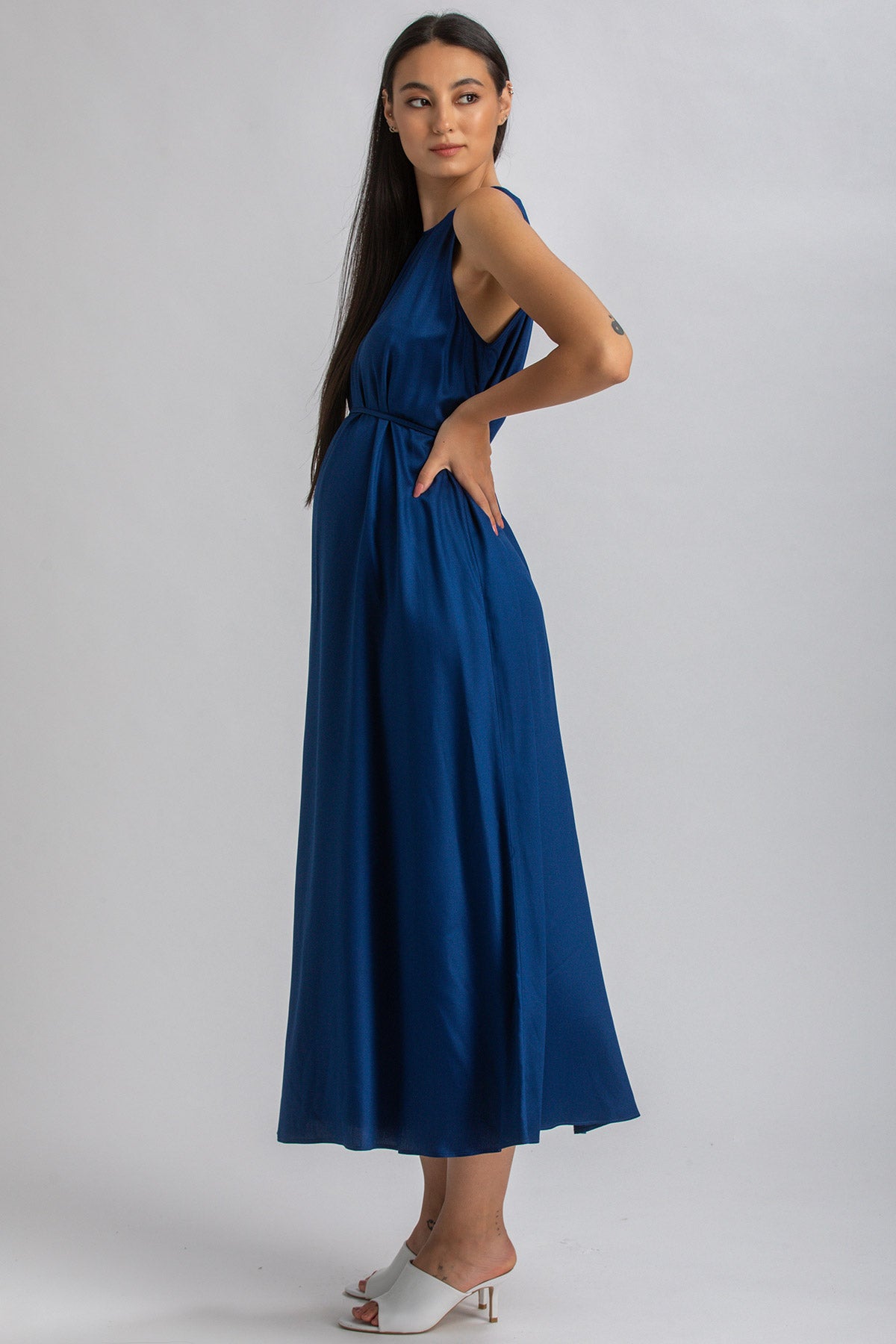 Donna incinta indossa abito premaman lungo blu elegante con sandali bianchi, sfondo neutro.
