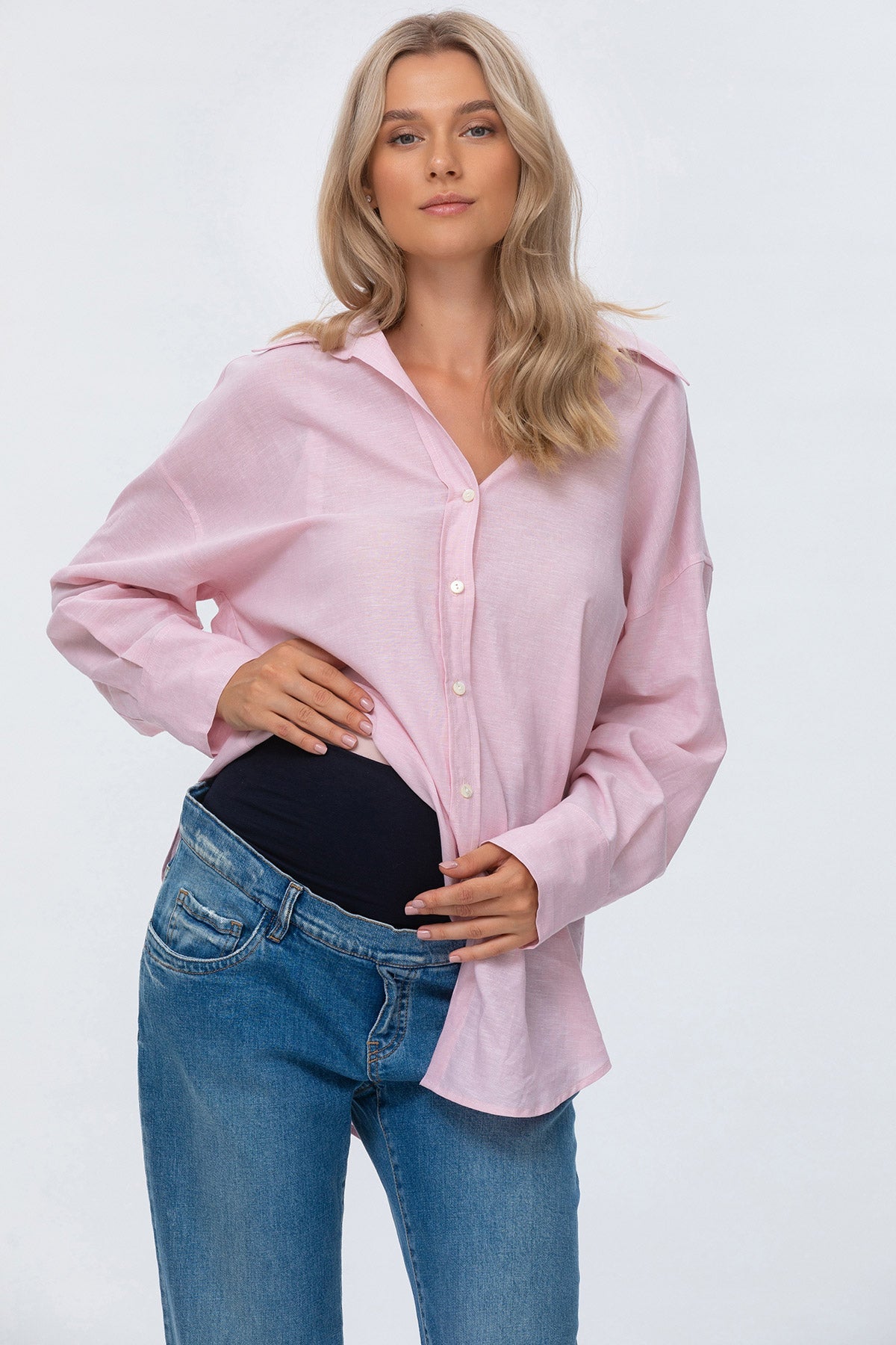 Donna incinta indossa camicia rosa premaman e jeans premaman a vita alta, fondo bianco.
