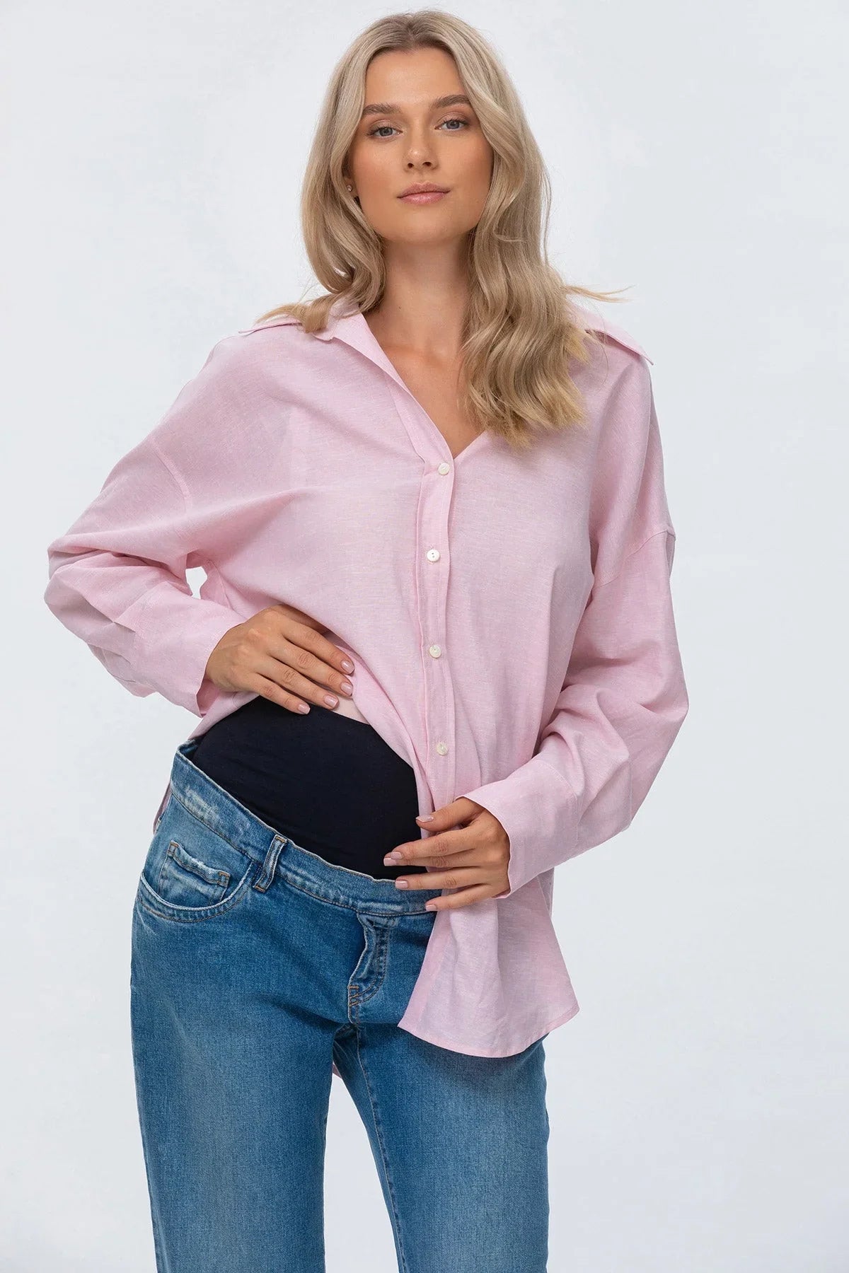 Donna incinta indossa camicia rosa premaman e jeans premaman a vita alta, fondo bianco.