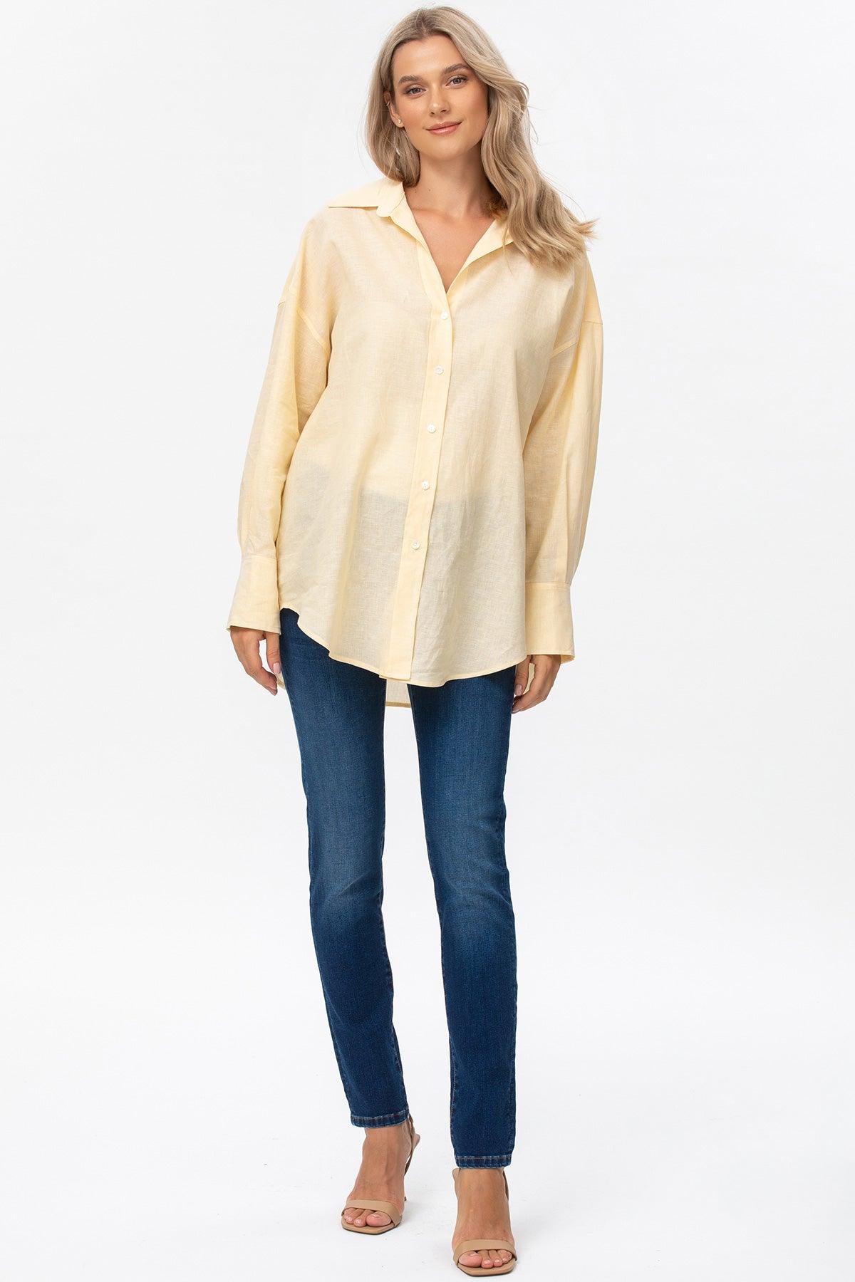 Donna con camicia premaman gialla a maniche lunghe e jeans aderenti, su sfondo bianco.