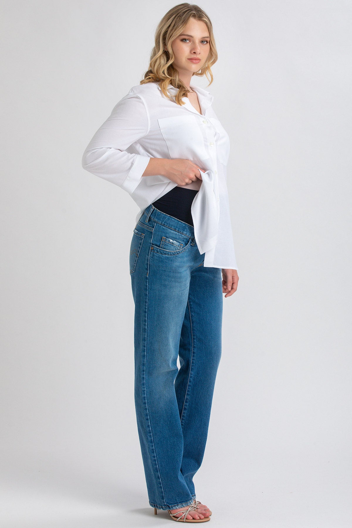 Donna incinta con camicia bianca e jeans premaman dritti in denim blu, su sfondo neutro.