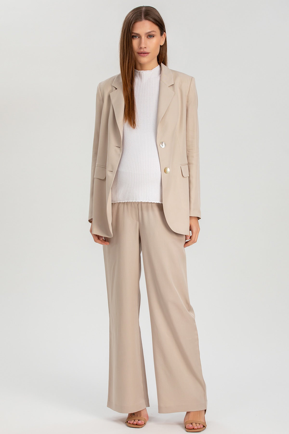 Donna incinta con completo premaman beige, blazer e pantaloni larghi, maglia bianca, sfondo neutro.