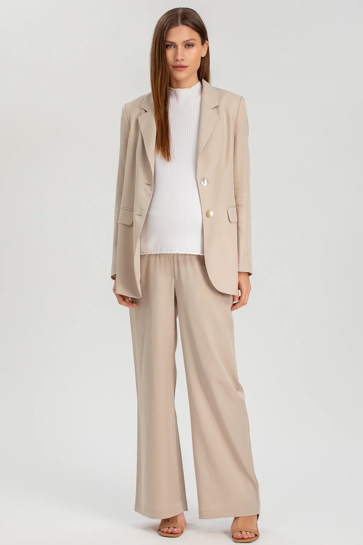 Donna incinta con completo premaman beige, blazer e pantaloni larghi, maglia bianca, sfondo neutro.