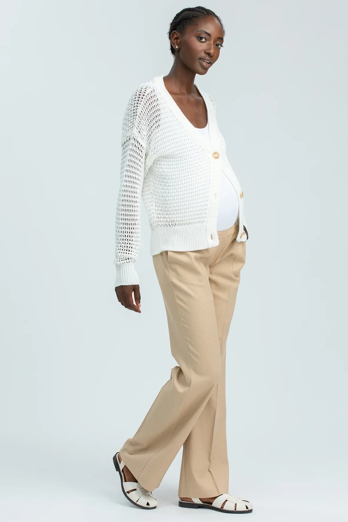 Modella incinta indossa cardigan premaman bianco traforato, pantaloni beige e sandali eleganti.