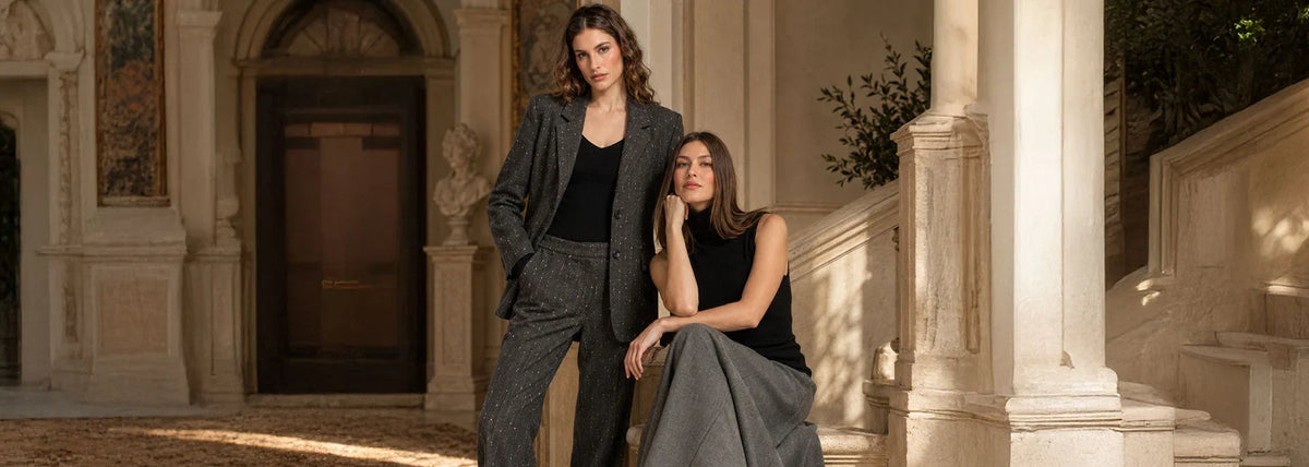 Due donne in una villa toscana: una indossa un completo sartoriale grigio scuro con dettagli luminosi, l'altra un top nero e una gonna lunga grigia.