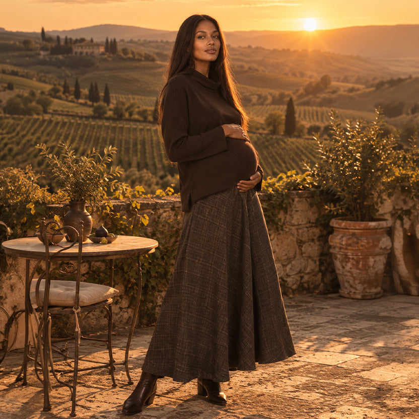 Donna incinta con maglione premaman marrone a collo alto e gonna midi a quadri, su una terrazza panoramica in Toscana al tramonto.