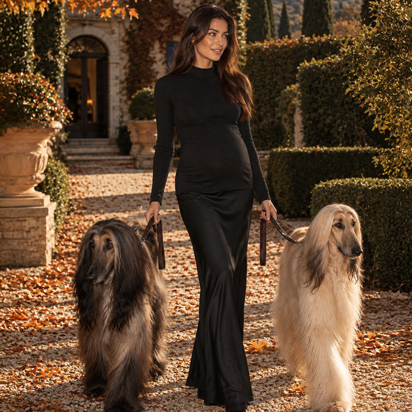 Donna incinta con abito premaman lungo nero aderente e maniche lunghe, mentre passeggia con due cani nel giardino autunnale di una villa toscana.