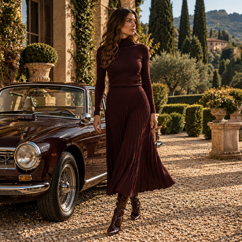 Donna con dolcevita bordeaux e gonna midi plissettata coordinata, accanto a un'auto d'epoca nel giardino di una villa toscana.