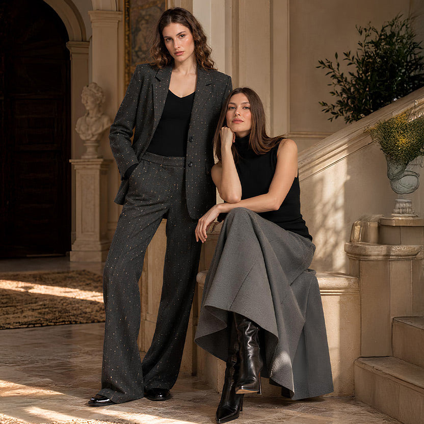 Due donne in una villa toscana: una indossa un completo sartoriale grigio scuro con dettagli luminosi, l'altra un top nero e una gonna lunga grigia.