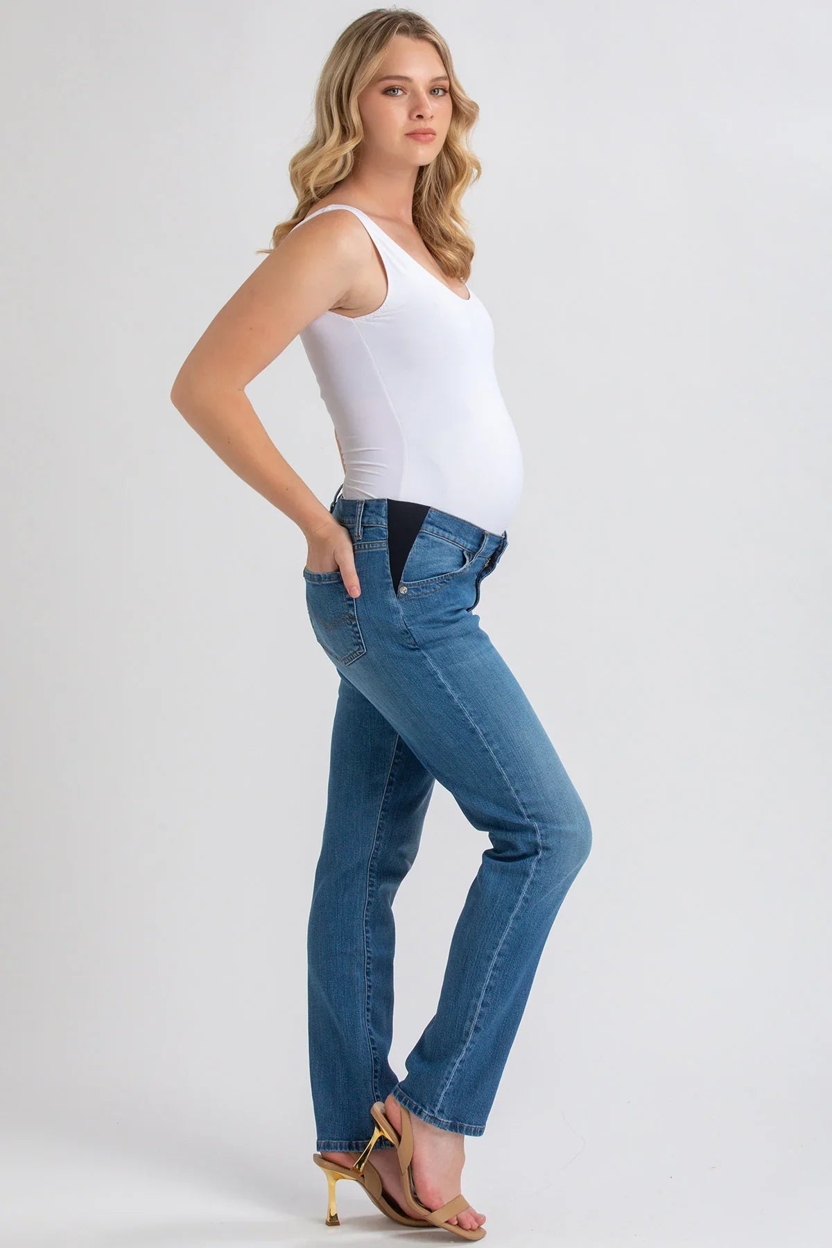 Donna incinta con jeans premaman a vita bassa e top bianco, posa su sfondo neutro