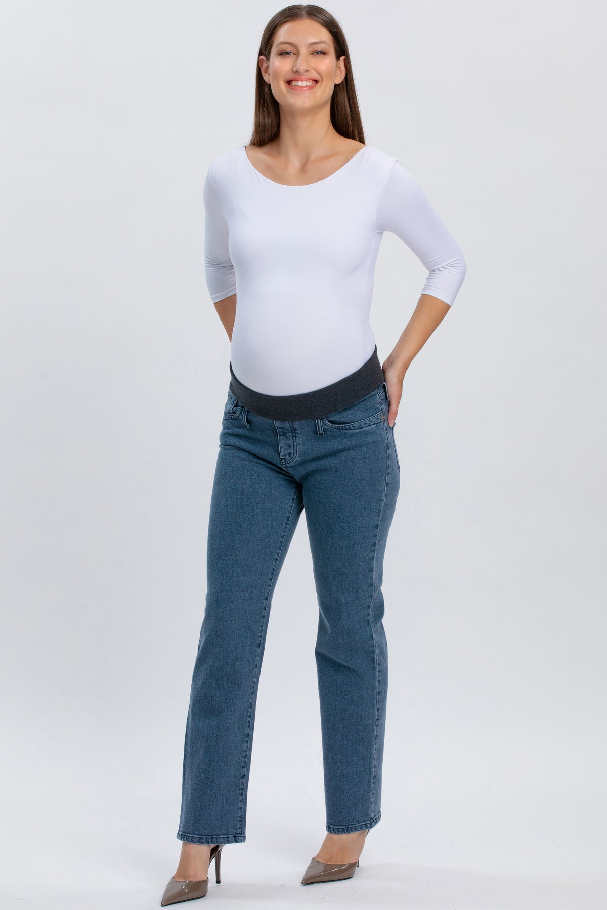Donna incinta con jeans premaman grigi a gamba dritta e maglia bianca, su sfondo neutro.