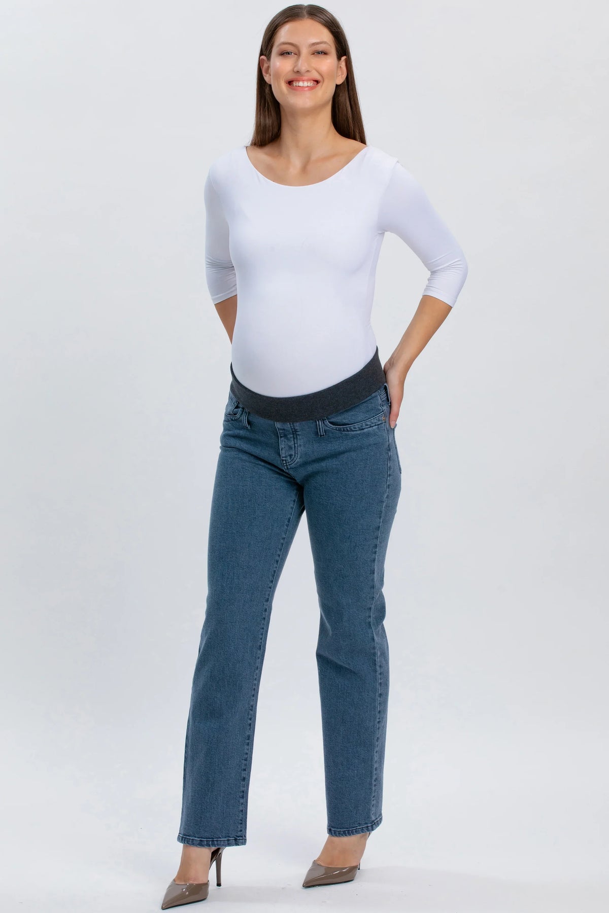 Donna incinta con jeans premaman grigi a gamba dritta e maglia bianca, su sfondo neutro.