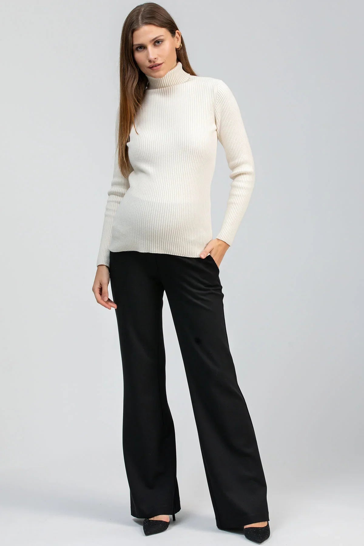 Modella indossa maglia premaman bianca a costine con collo alto e pantaloni neri a gamba larga.