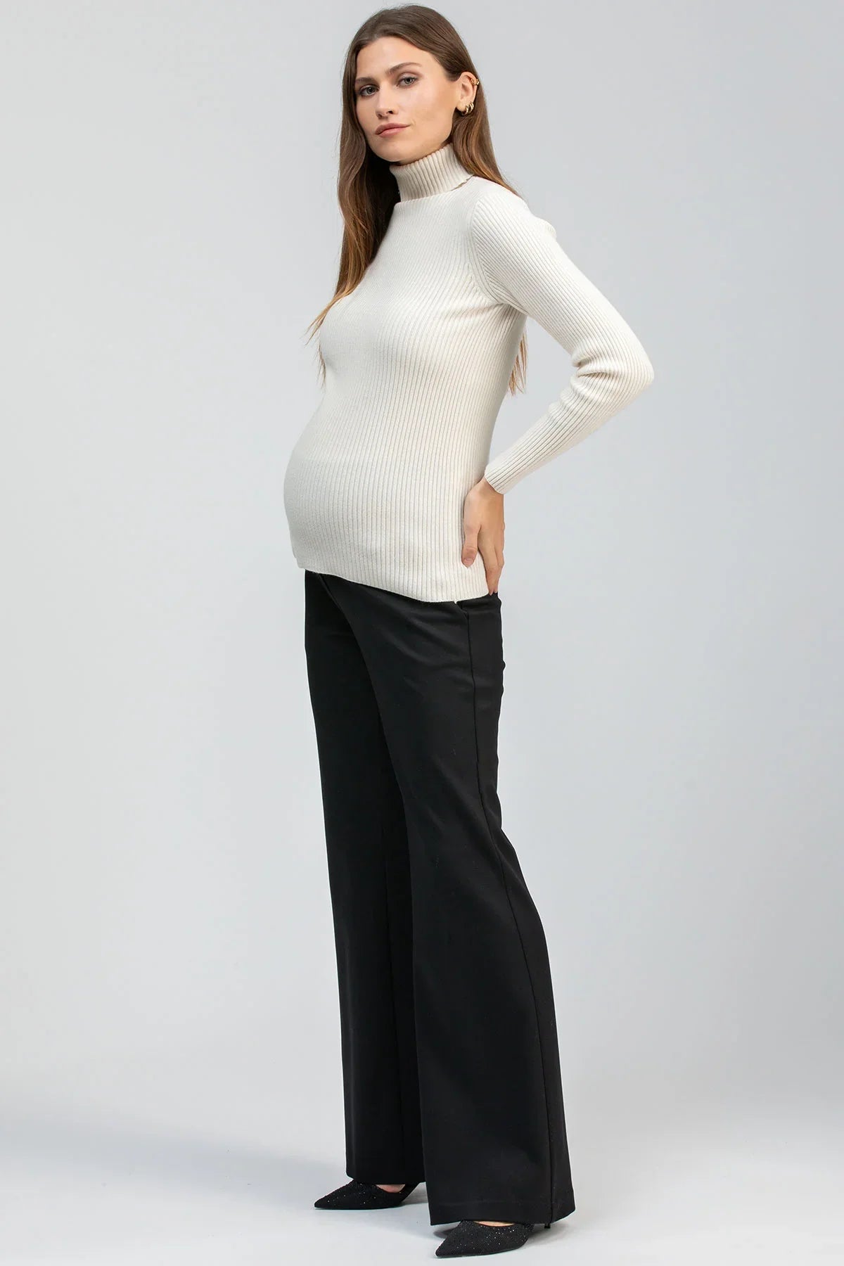 Modella indossa maglia premaman bianca a costine con collo alto e pantaloni neri a gamba larga.