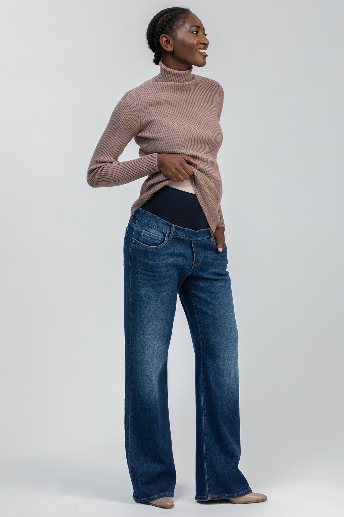 Modella indossa jeans premaman a zampa e maglione a collo alto beige su sfondo neutro.