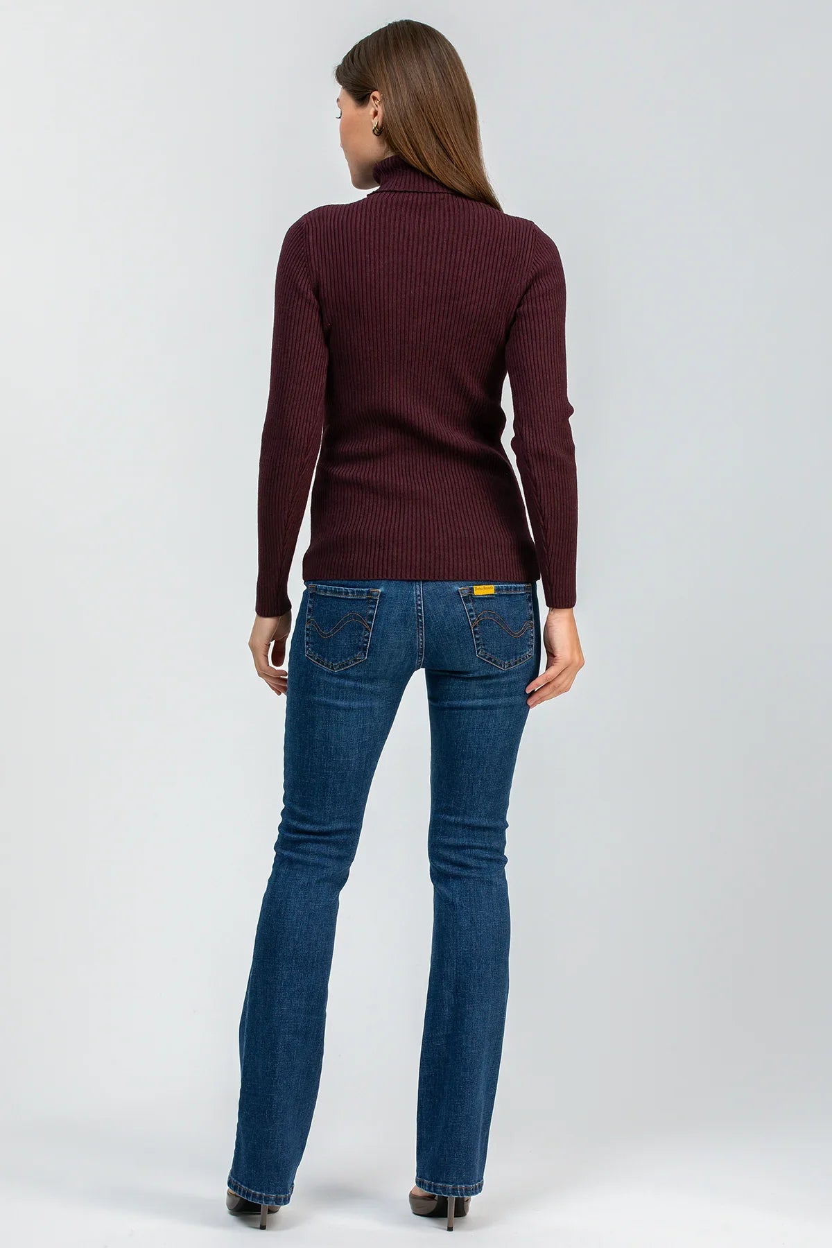 Donna di spalle con maglione bordeaux e jeans premaman bootcut blu, su sfondo neutro