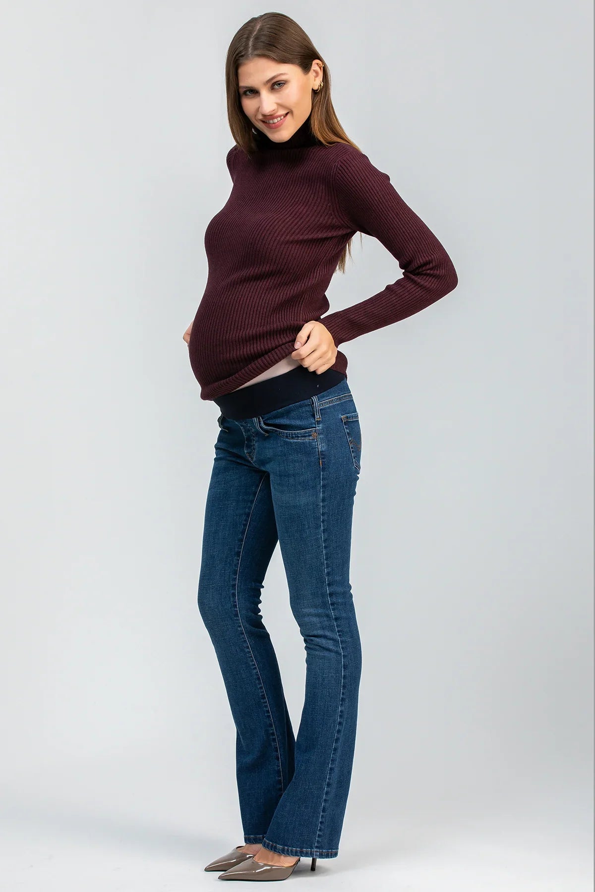 Modella incinta indossa jeans premaman bootcut blu e maglione bordeaux, fondo neutro.