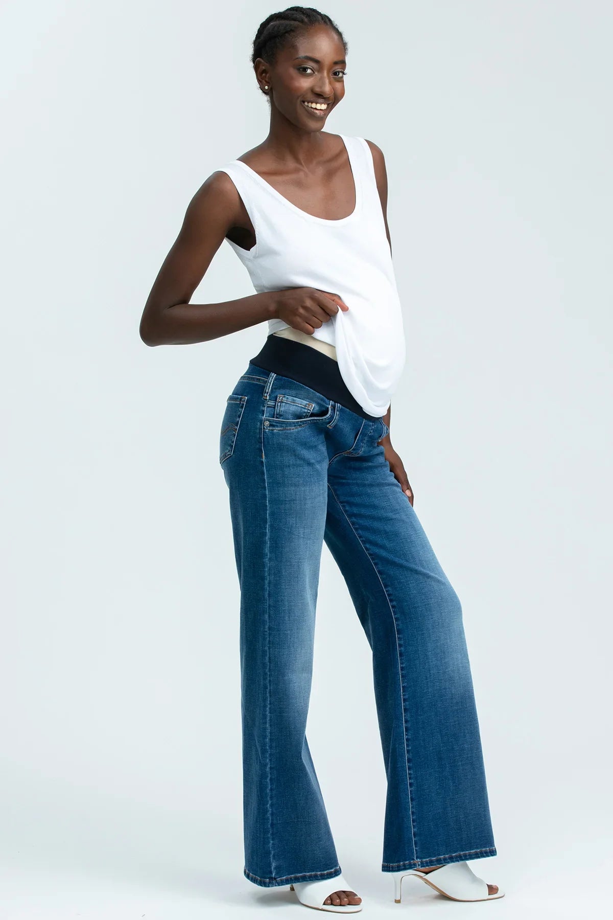 Donna incinta indossa jeans premaman a gamba larga in denim blu e top bianco, fondo bianco.