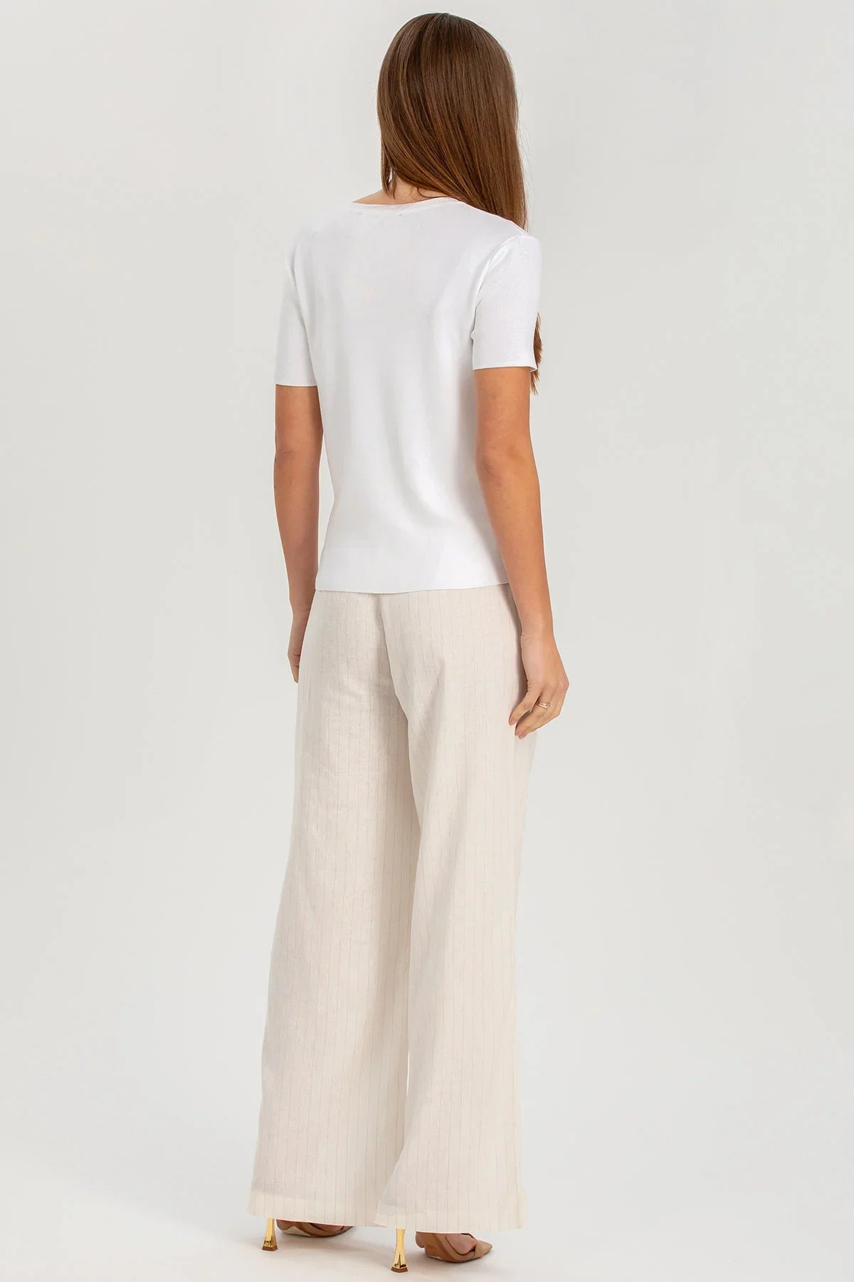 Donna di spalle con maglia bianca e pantaloni premaman larghi color panna, fondo neutro