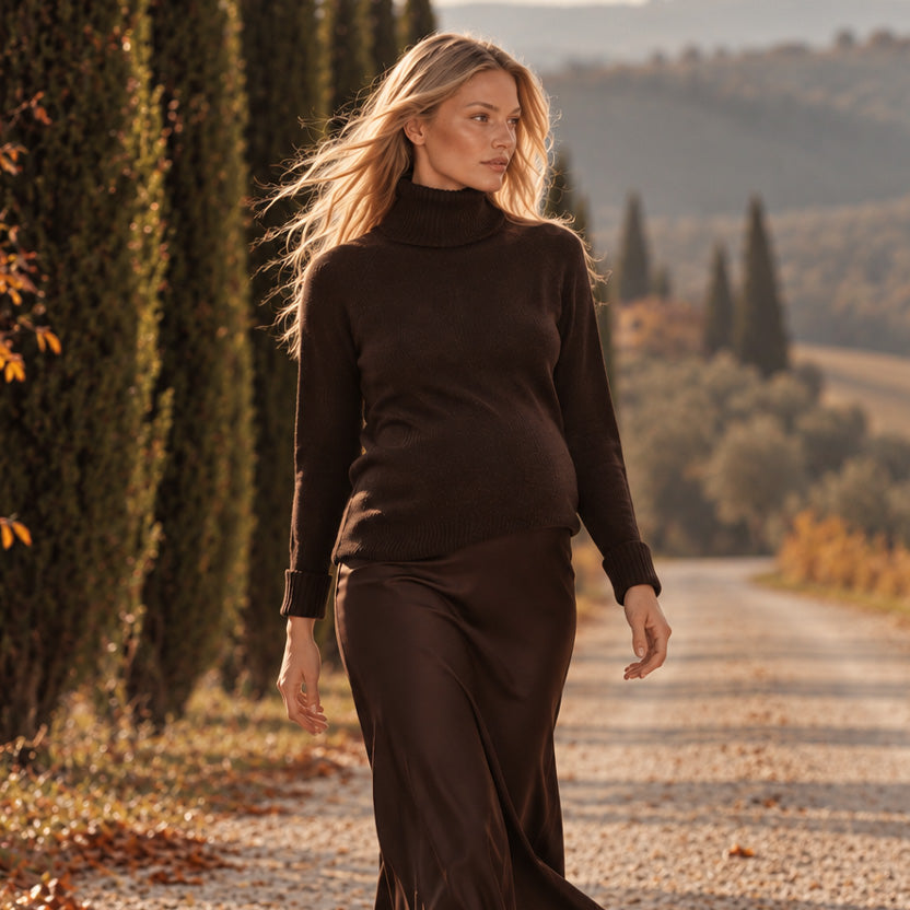 Donna incinta bionda con completo premaman marrone composto da maglione a collo alto e gonna lunga coordinata, mentre passeggia lungo un viale alberato in Toscana.