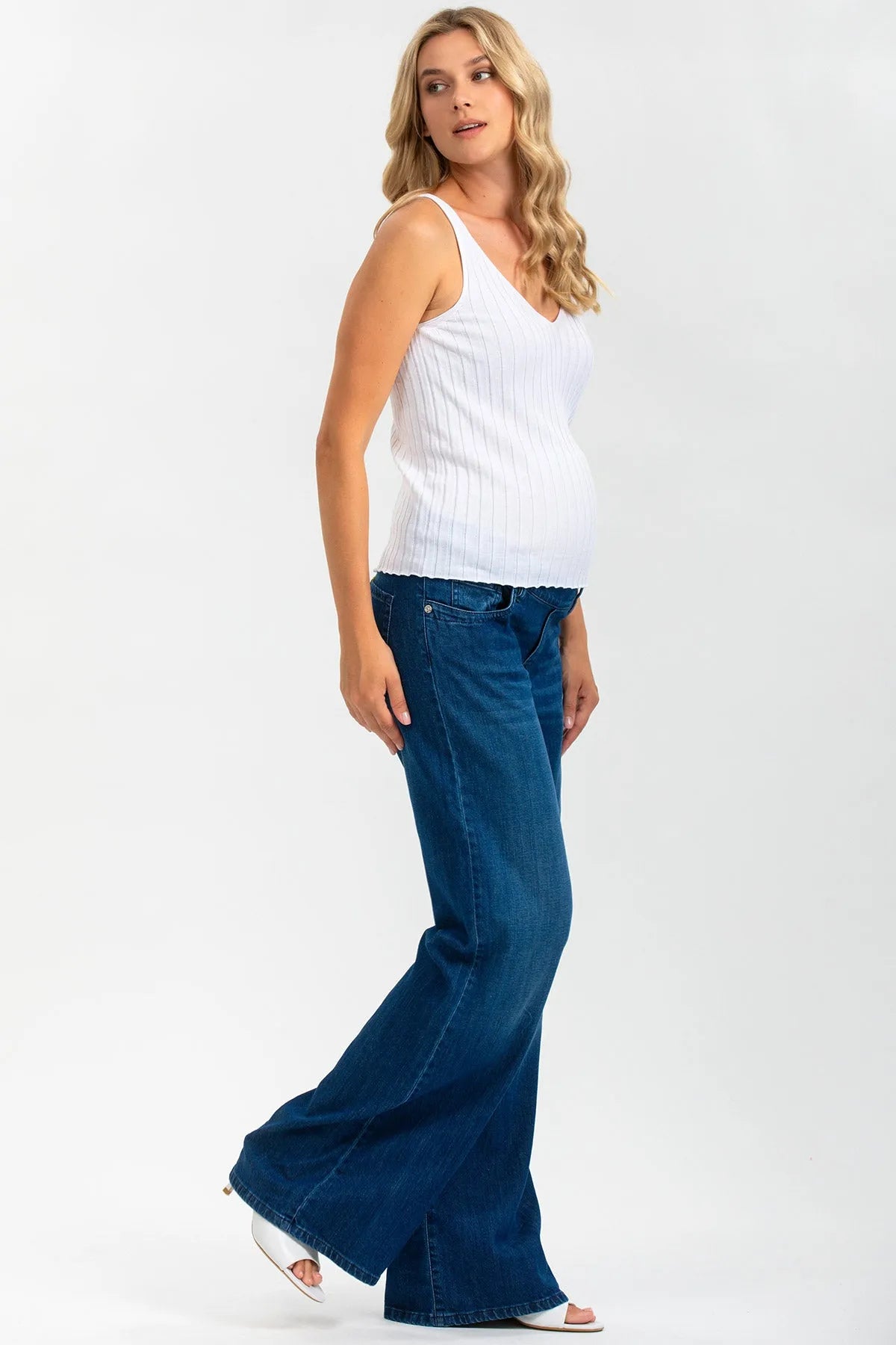 Donna incinta con top bianco senza maniche e jeans premaman a zampa su sfondo chiaro