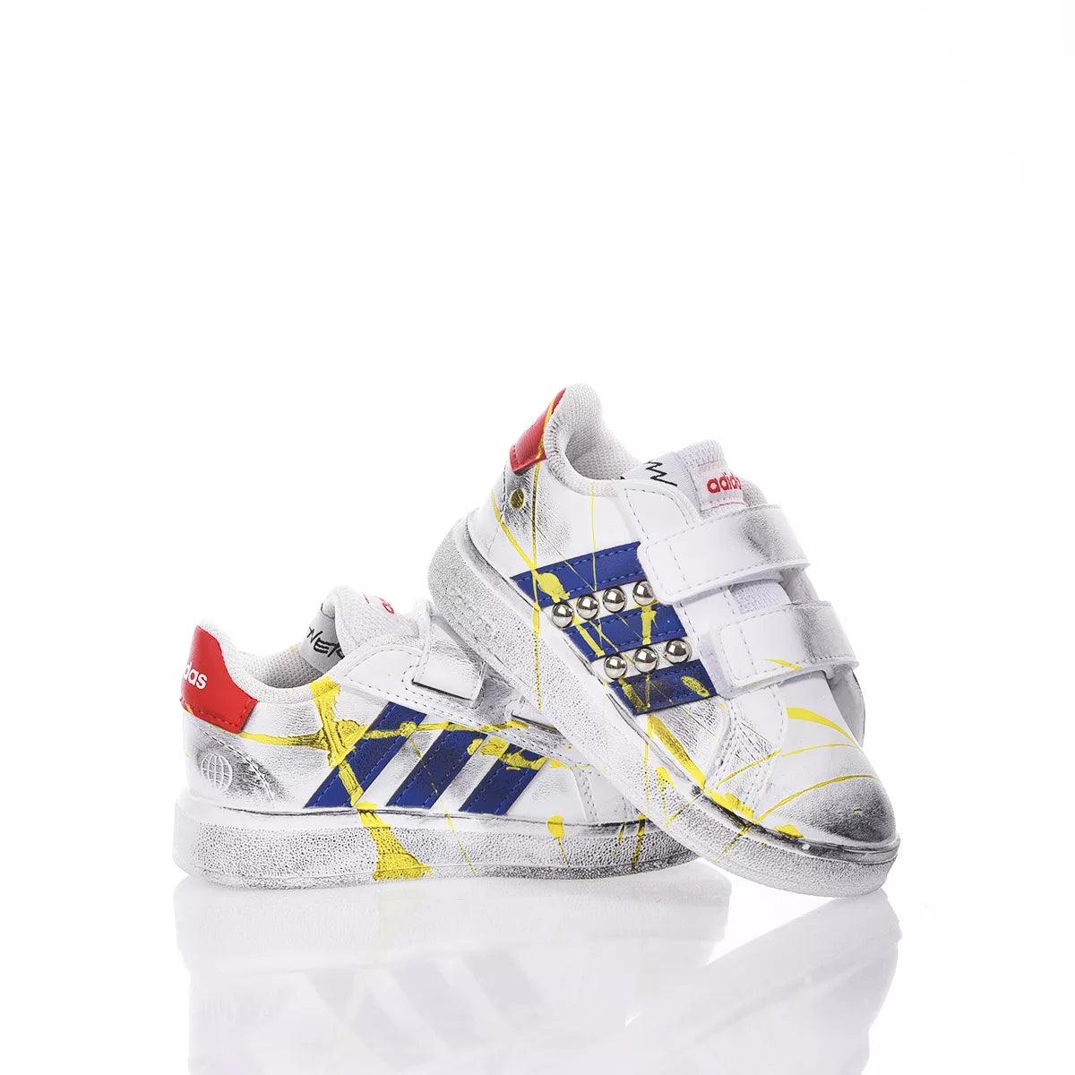 Sneakers bianche con dettagli blu e gialli, borchie e chiusura a strappo su sfondo bianco.