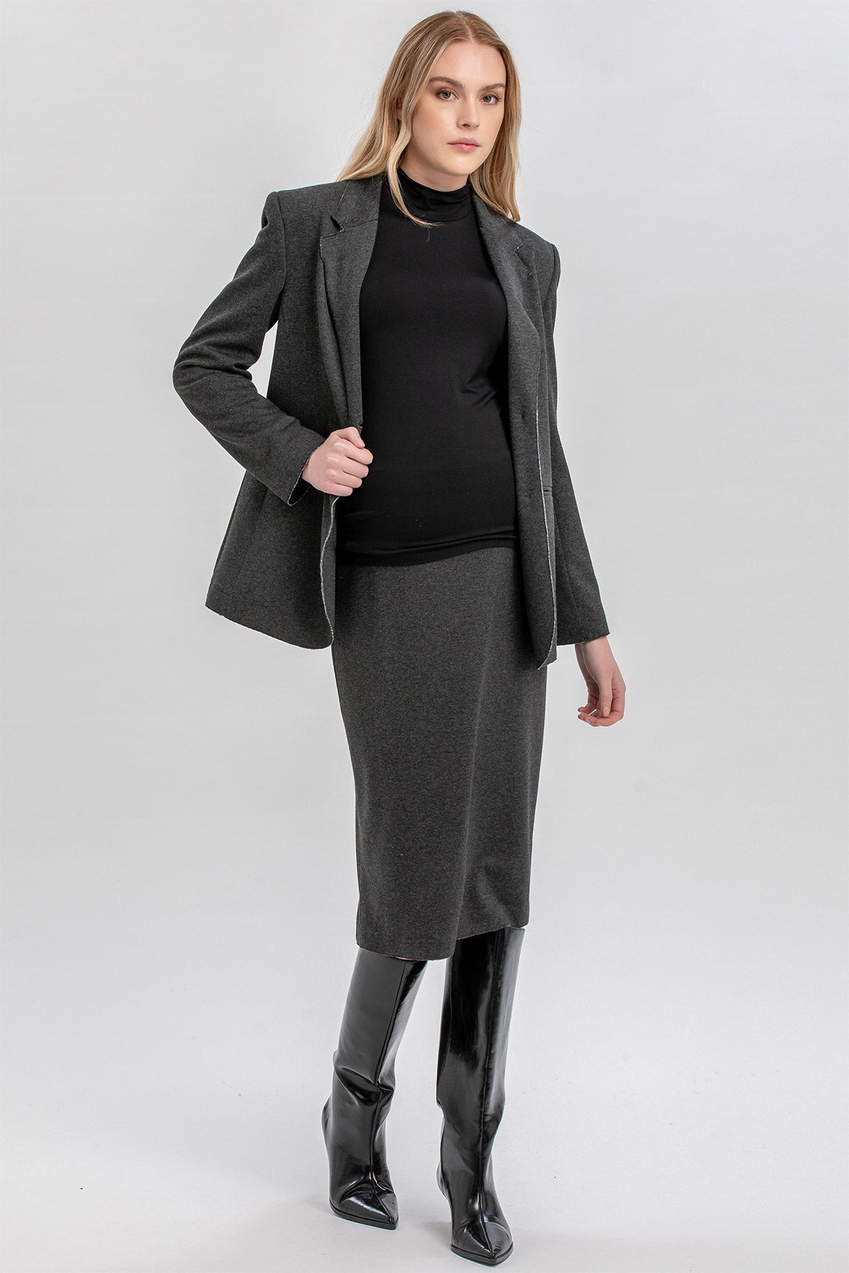 Donna incinta con giacca premaman grigia, dolcevita nero e stivali alti neri, look elegante