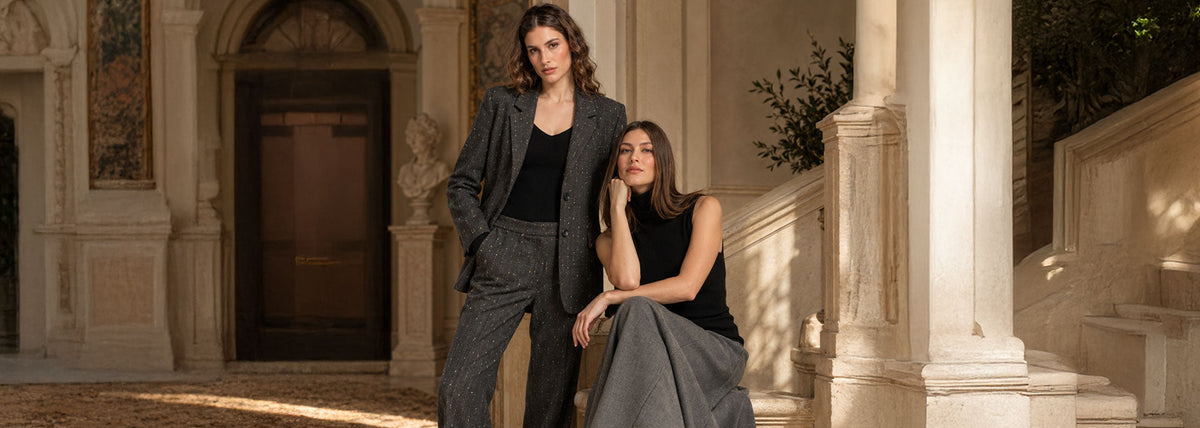 Due donne in una villa toscana: una indossa un completo sartoriale grigio scuro con dettagli luminosi, l'altra un top nero e una gonna lunga grigia.