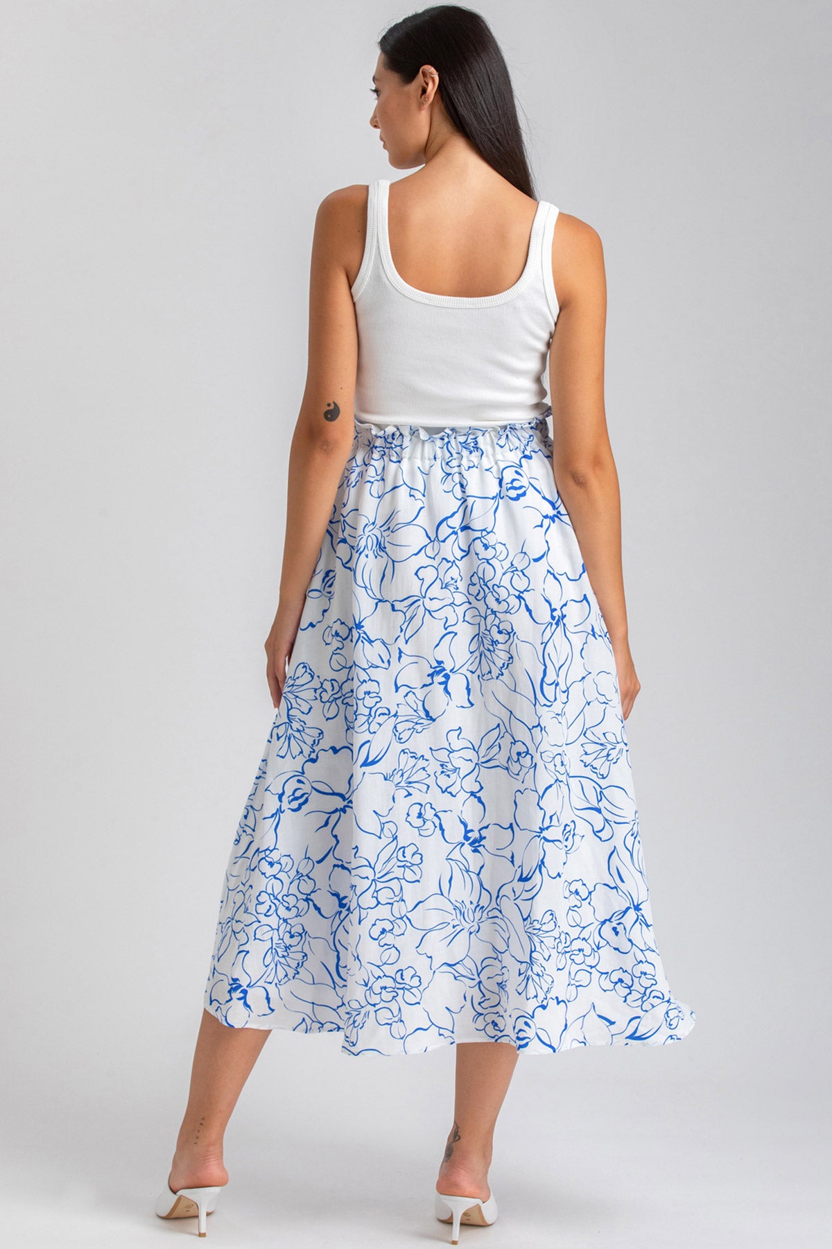 Donna di spalle con top bianco e gonna premaman lunga bianca a fiori blu, su sfondo neutro.