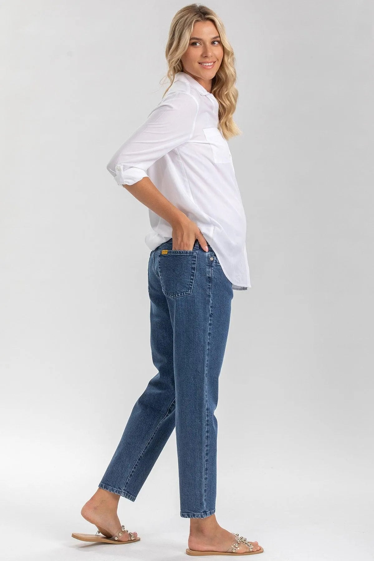 Donna incinta con camicia bianca premaman e jeans maternità blu, fondo neutro.