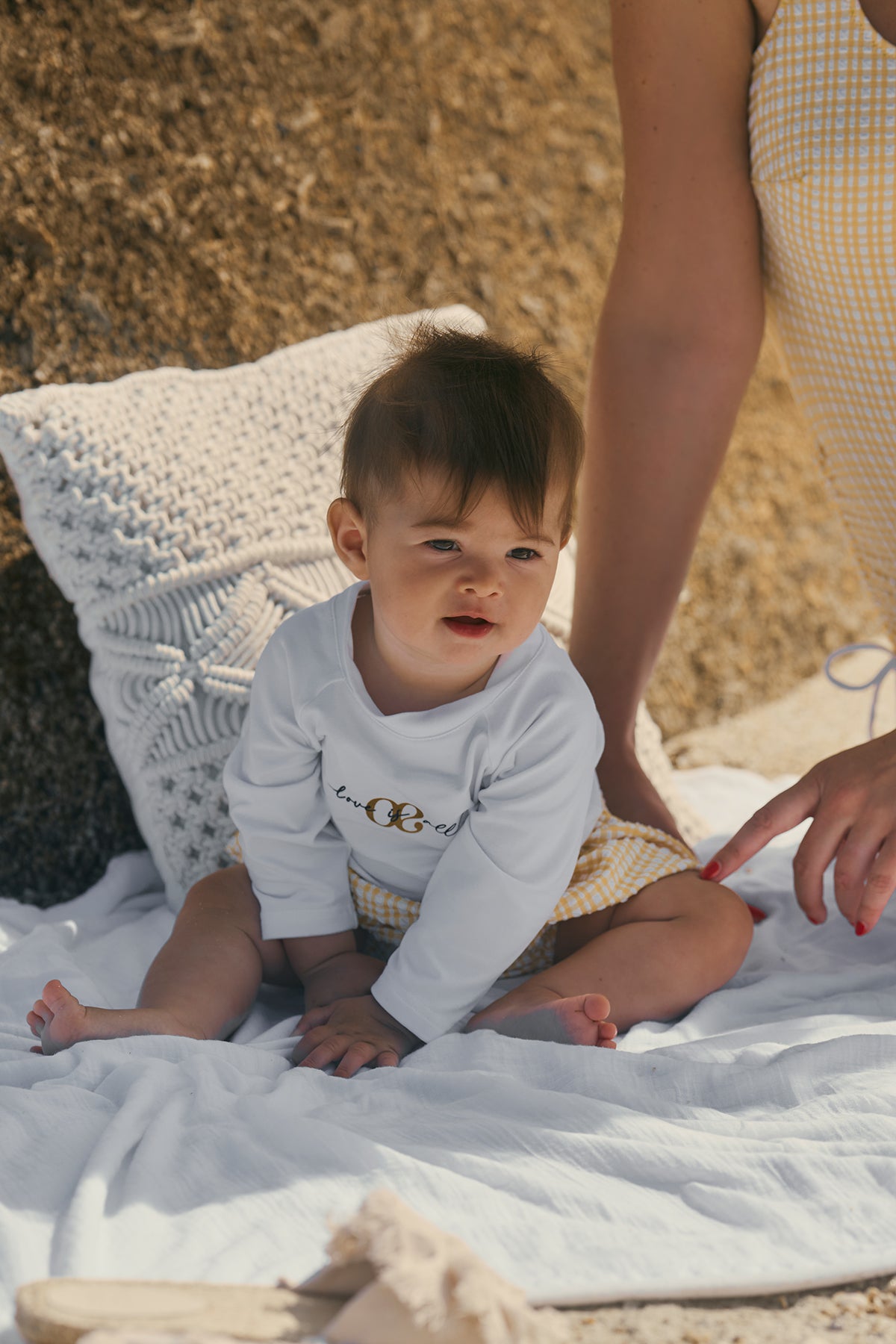 Bambino seduto su coperta bianca all'aperto, con maglia bianca e pantaloncini gialli a quadretti.