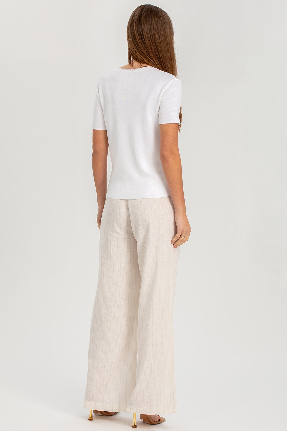 Donna di spalle con maglia bianca e pantaloni premaman larghi color panna, fondo neutro