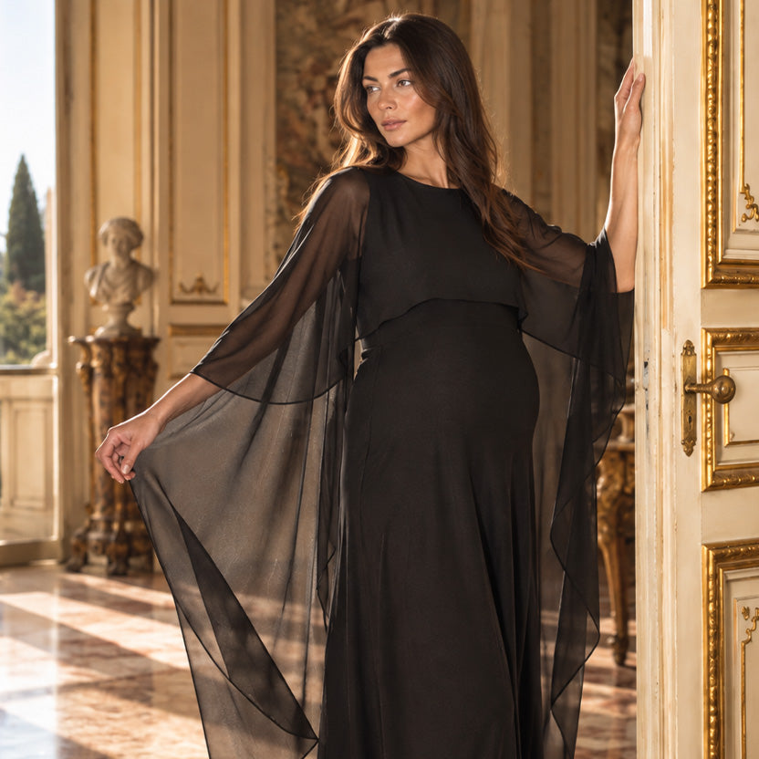 Donna incinta con vestito premaman elegante nero lungo, caratterizzato da ampie maniche a mantella in tessuto trasparente, in una villa toscana.
